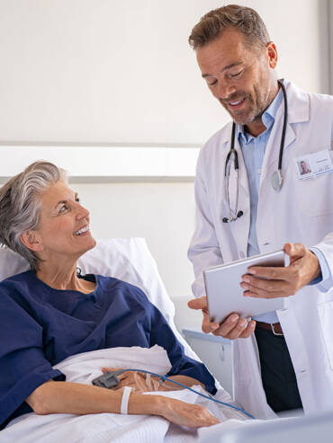Doctor in white coat consulting smiling female patient in bright hospital room with medical equipment and digital tablet.