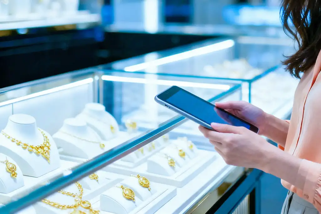 Asian woman in peach blouse holding a tablet in a bright, modern luxury jewelry store with illuminated gold and white displays.