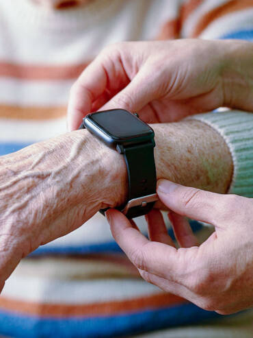 Caregiver in green scrubs adjusting black smart watch on elderly wrist with visible veins.