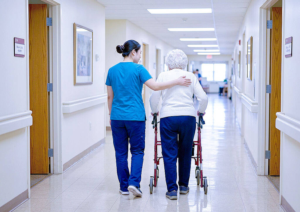 East Asian caregiver in teal scrubs assists elderly woman with walker in bright care home corridor.