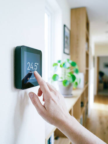 Hand tapping sleek black touchscreen thermostat displaying 24.5° on cream wall.