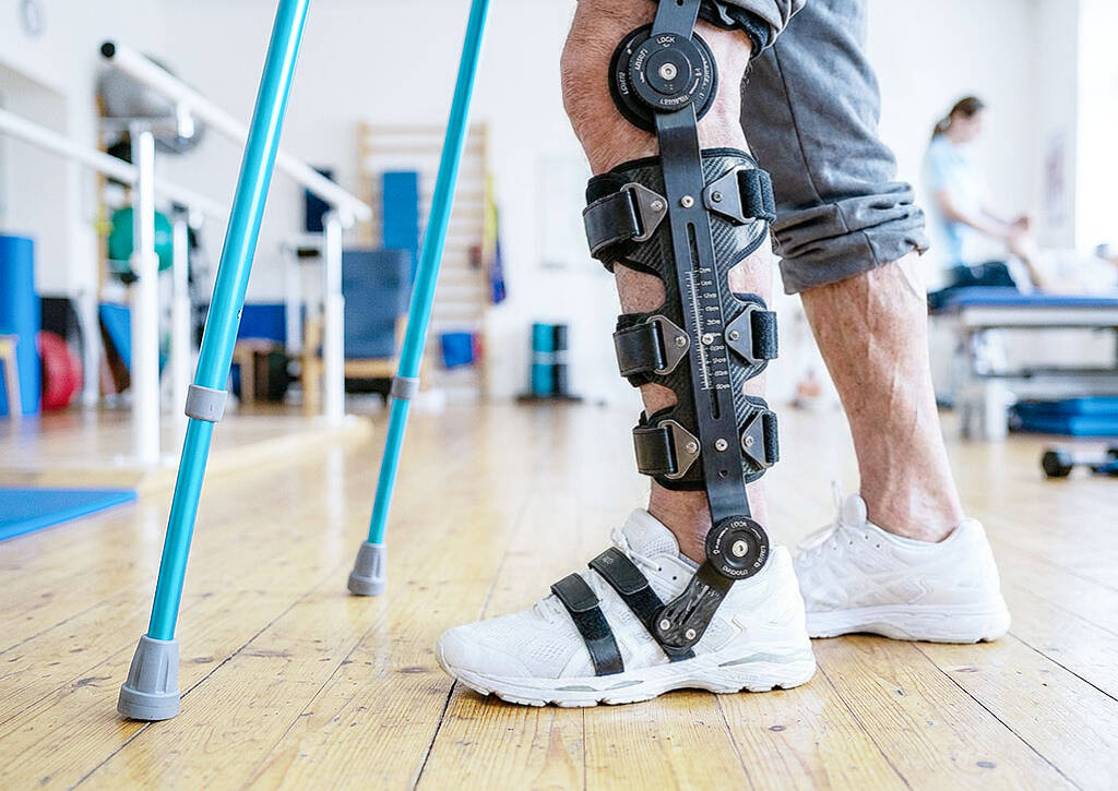 Close-up of black mechanical leg brace with buckles on denim jeans and blue sneakers, turquoise crutches nearby on wood floor.
